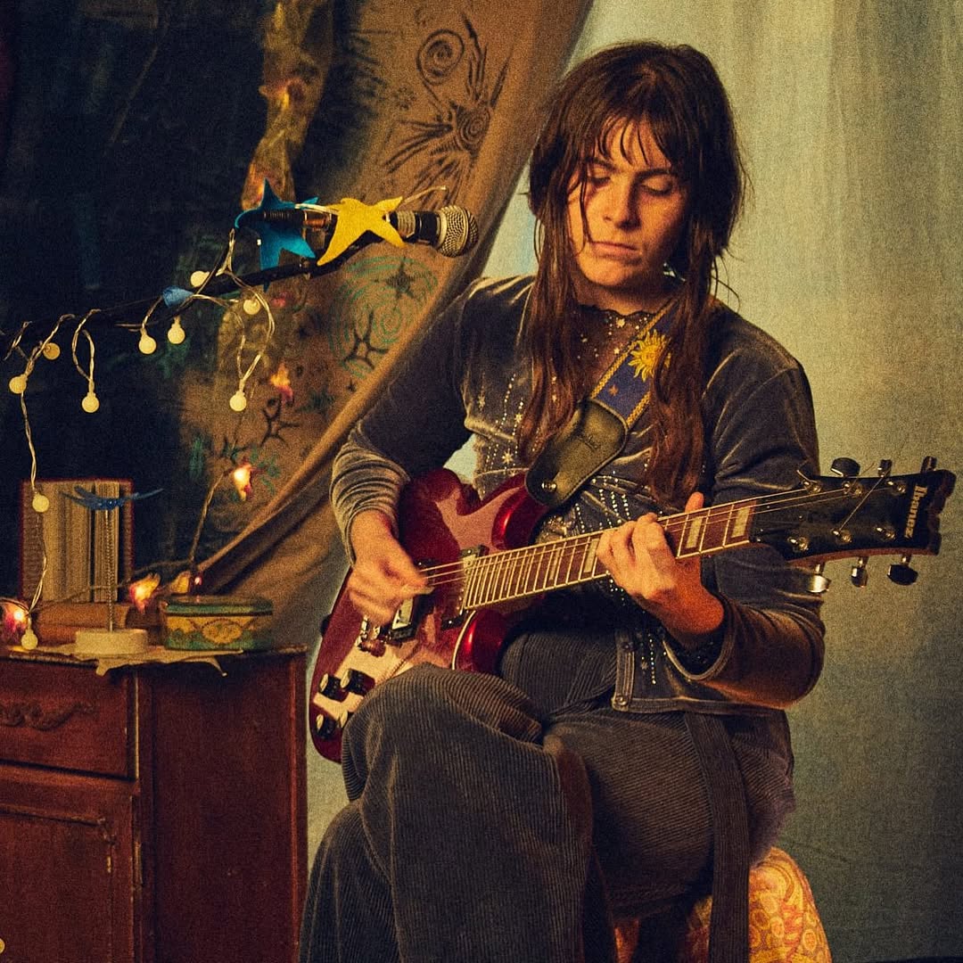 Celeste sitting on a stool strumming a guitar in front of a microphone