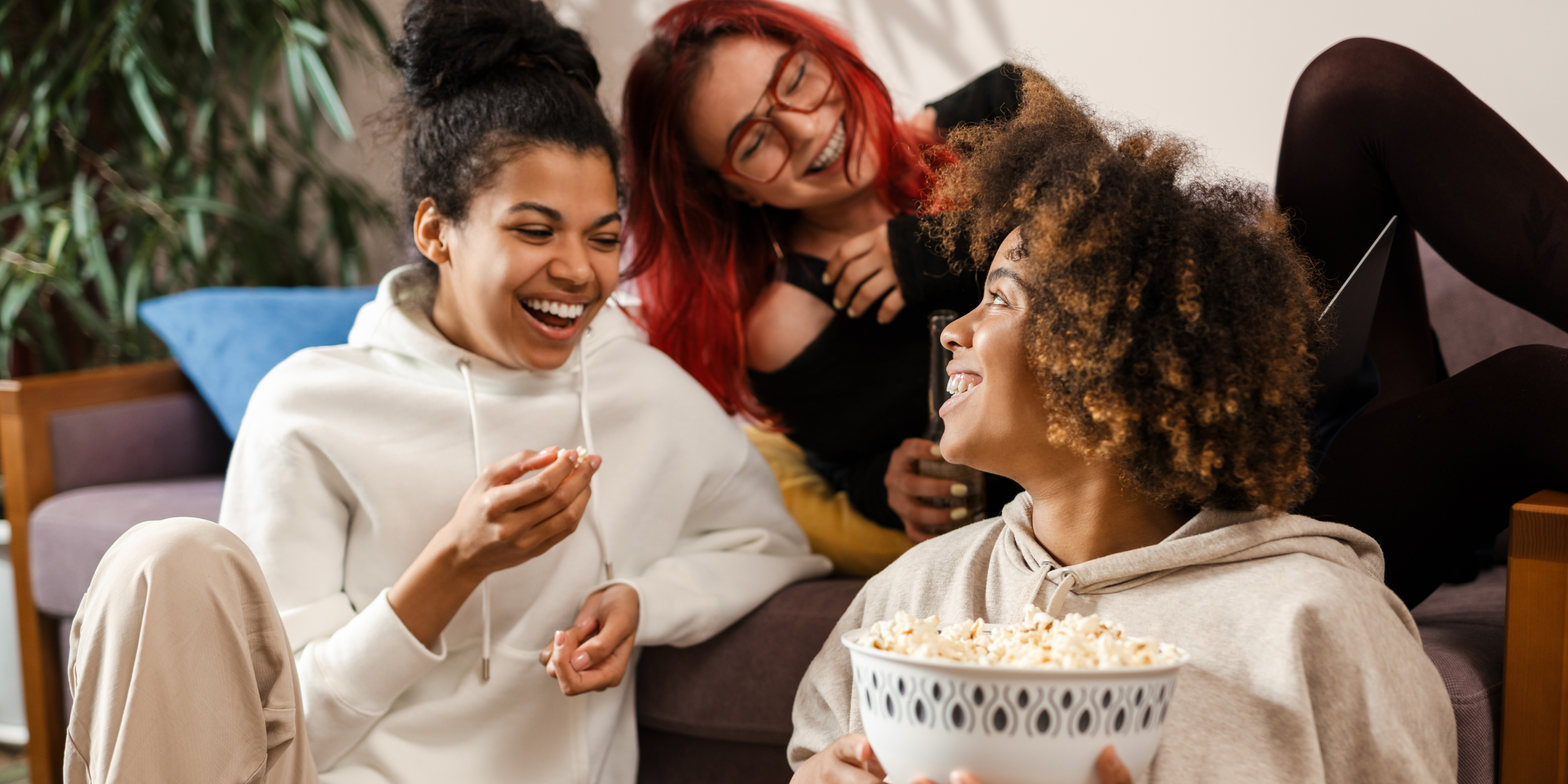 3 young women sitting on a couch laughing together eating popcorn