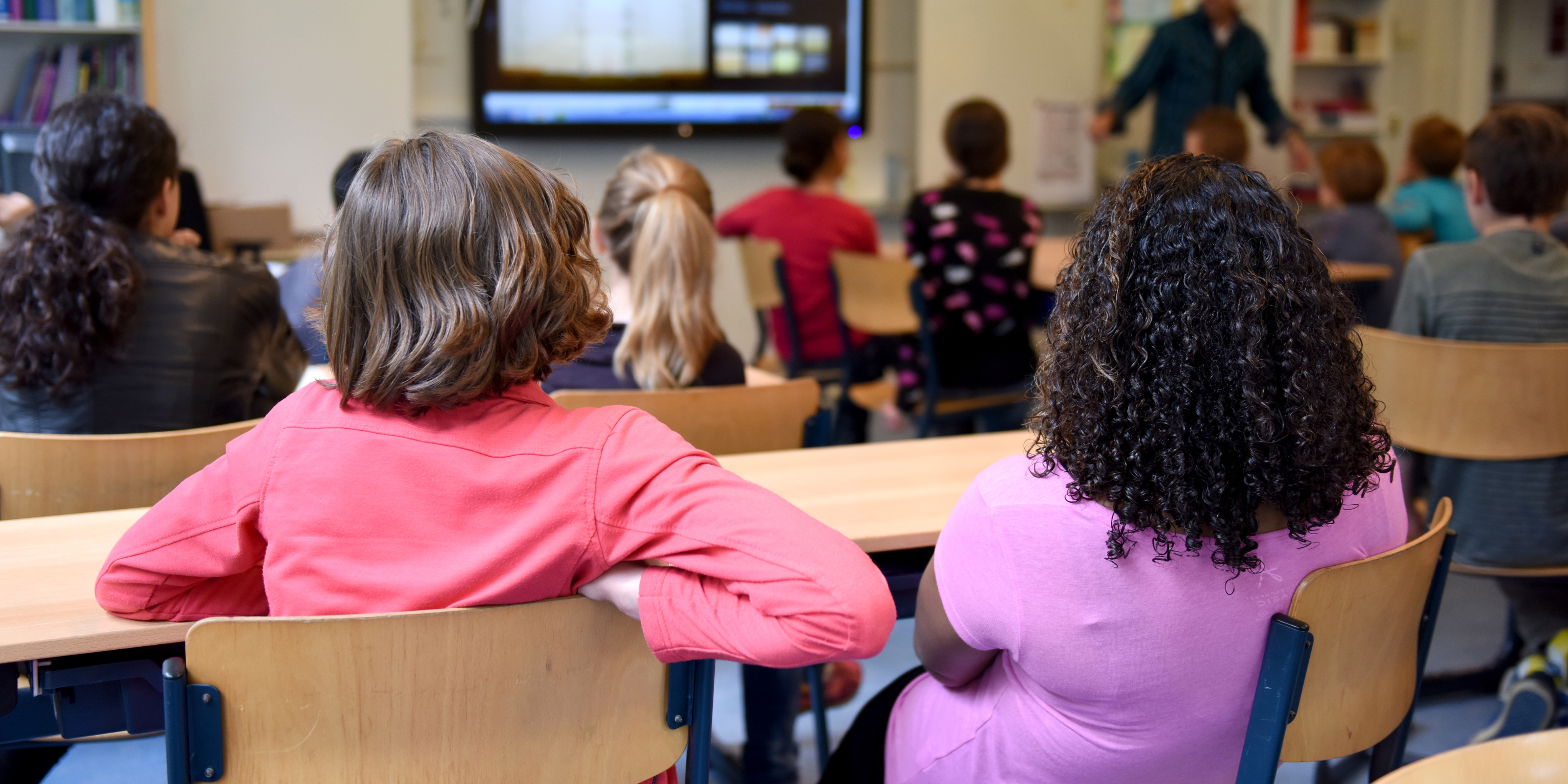 the backs of children sitting in a classroom listening to their teacher 