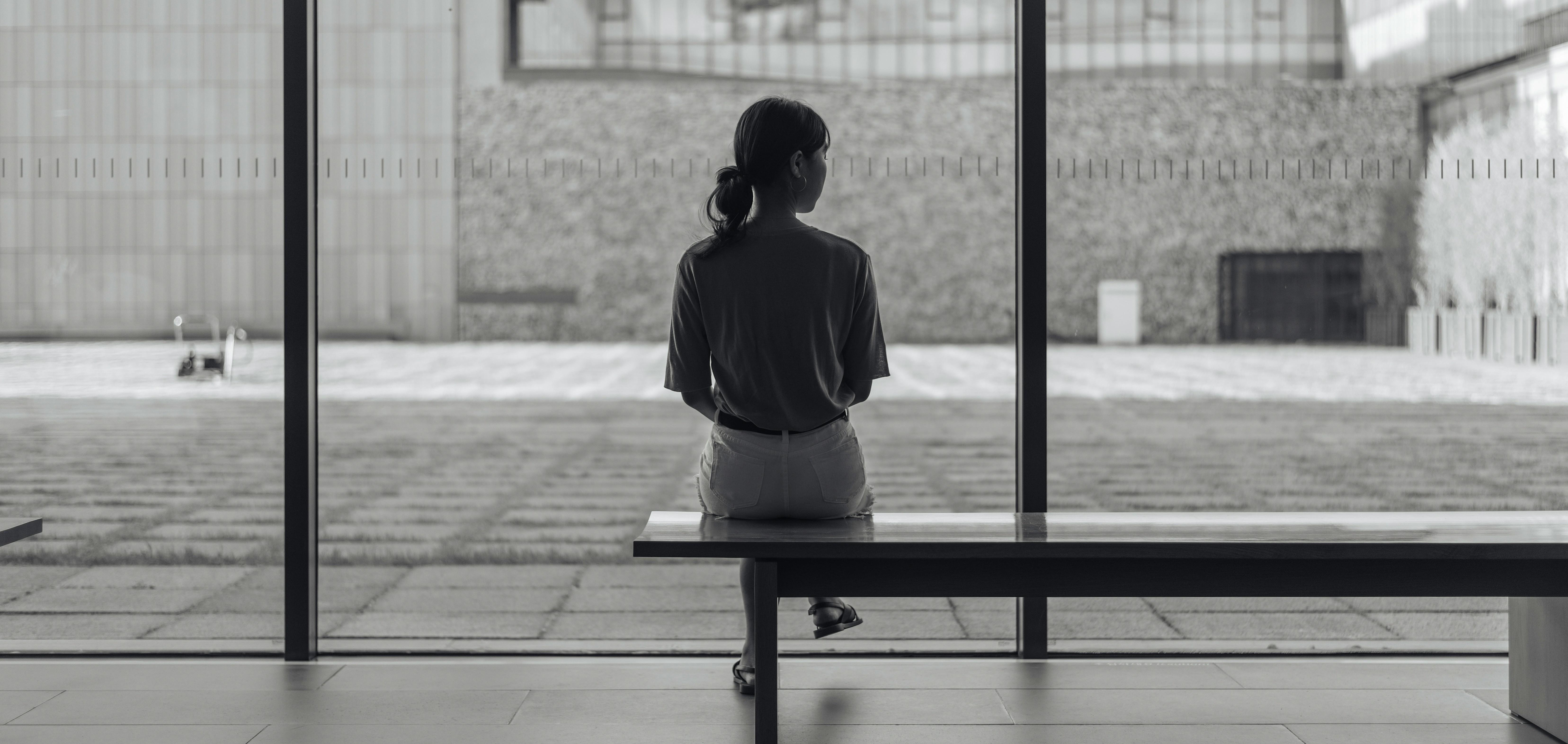 a black and white image of a young woman sitting by herself on a bench looking out large windows 