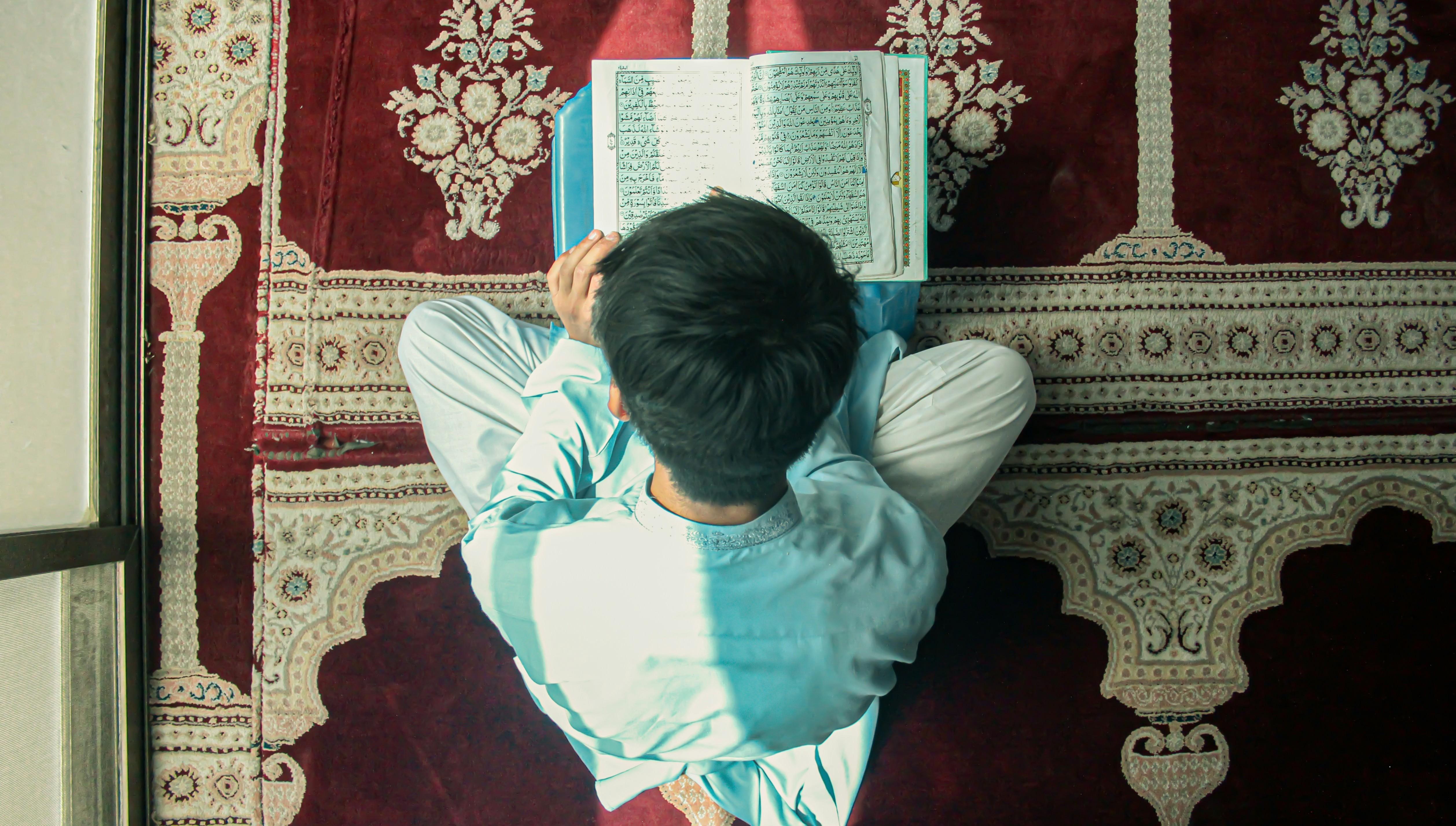 a young boy sitting on a prayer rug reading the Quran 