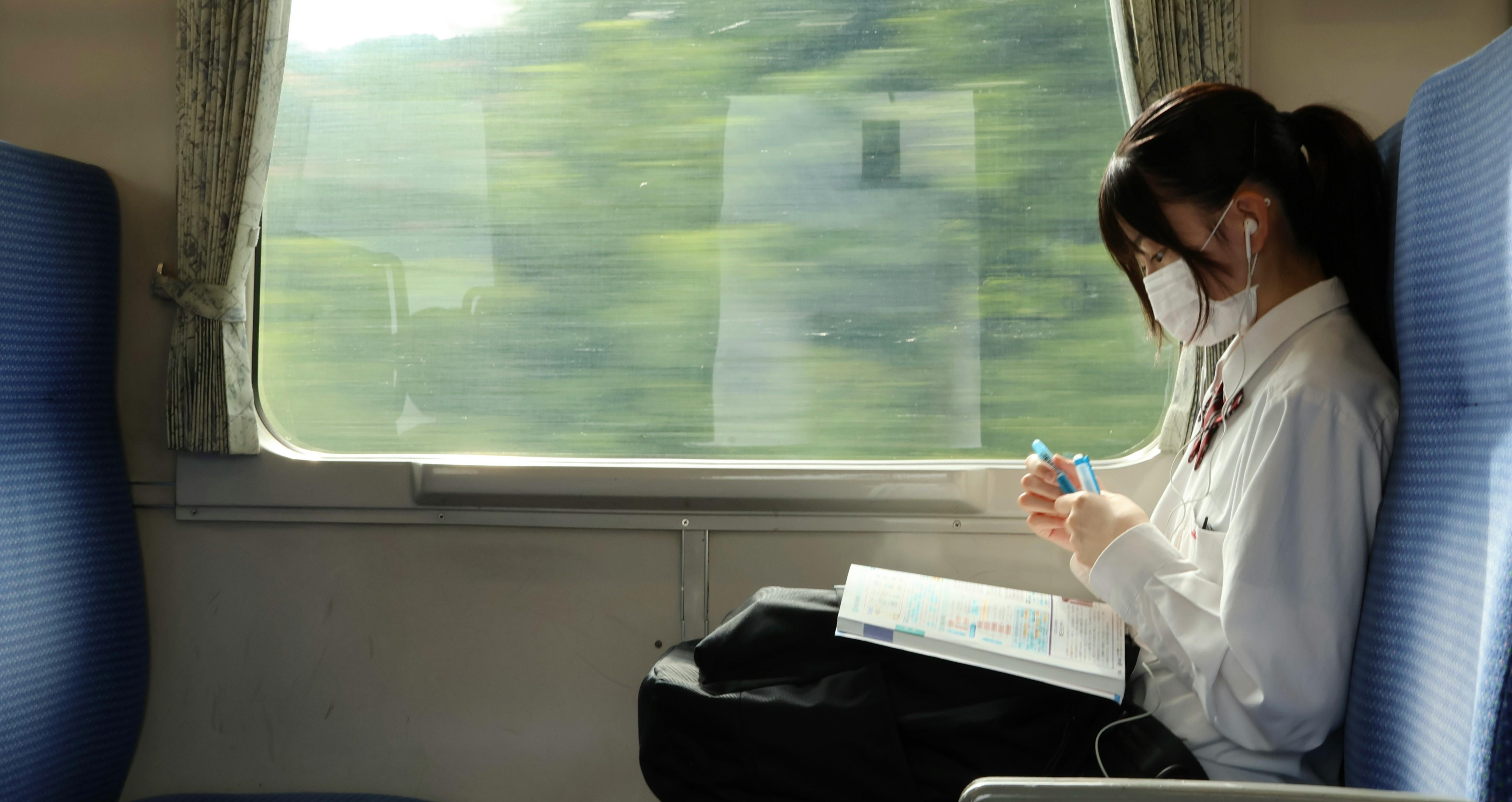 young woman sitting on the train with headphones and a mask on working in a book