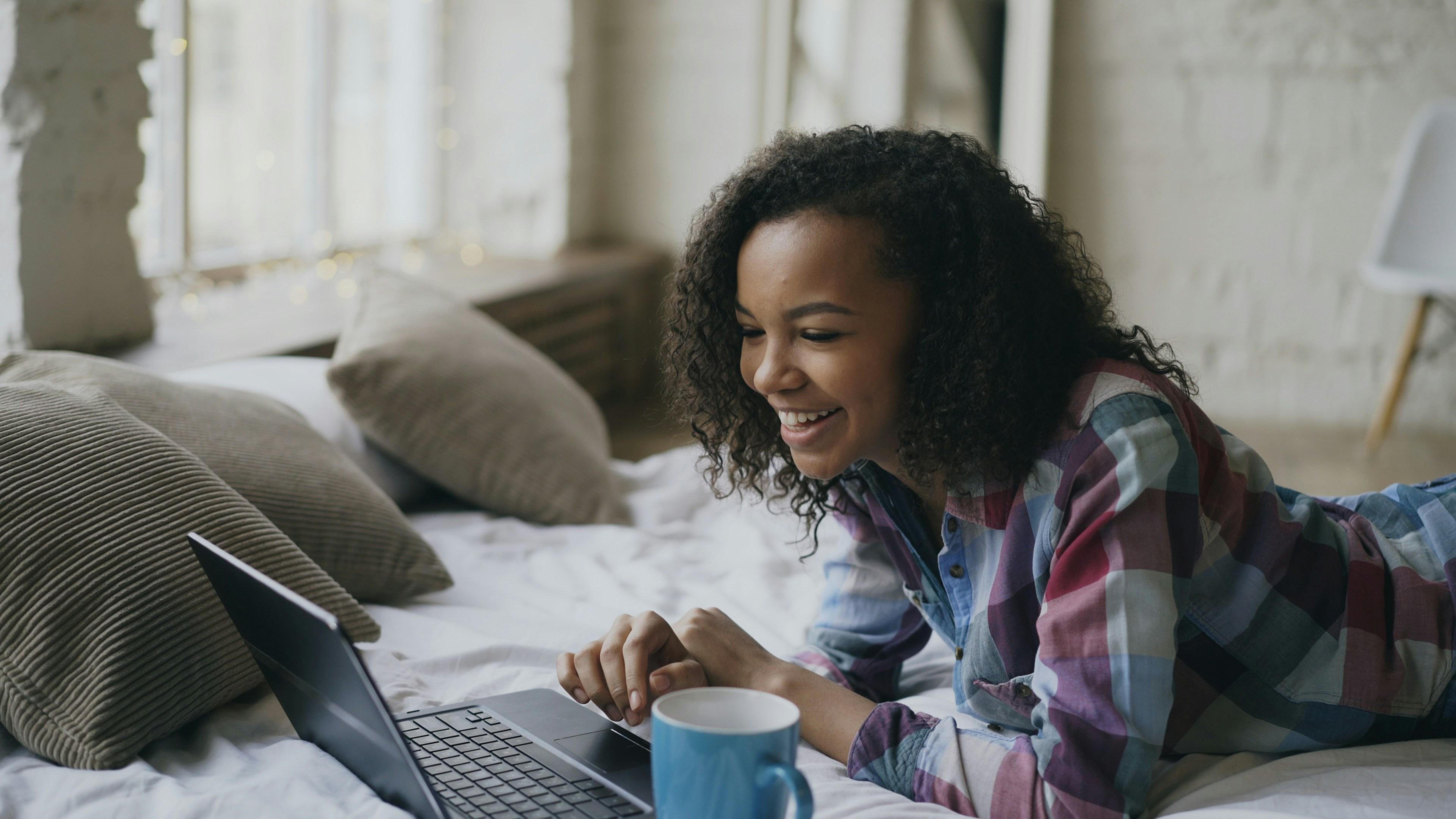 young girl lying on her bed typing on her laptop and smiling