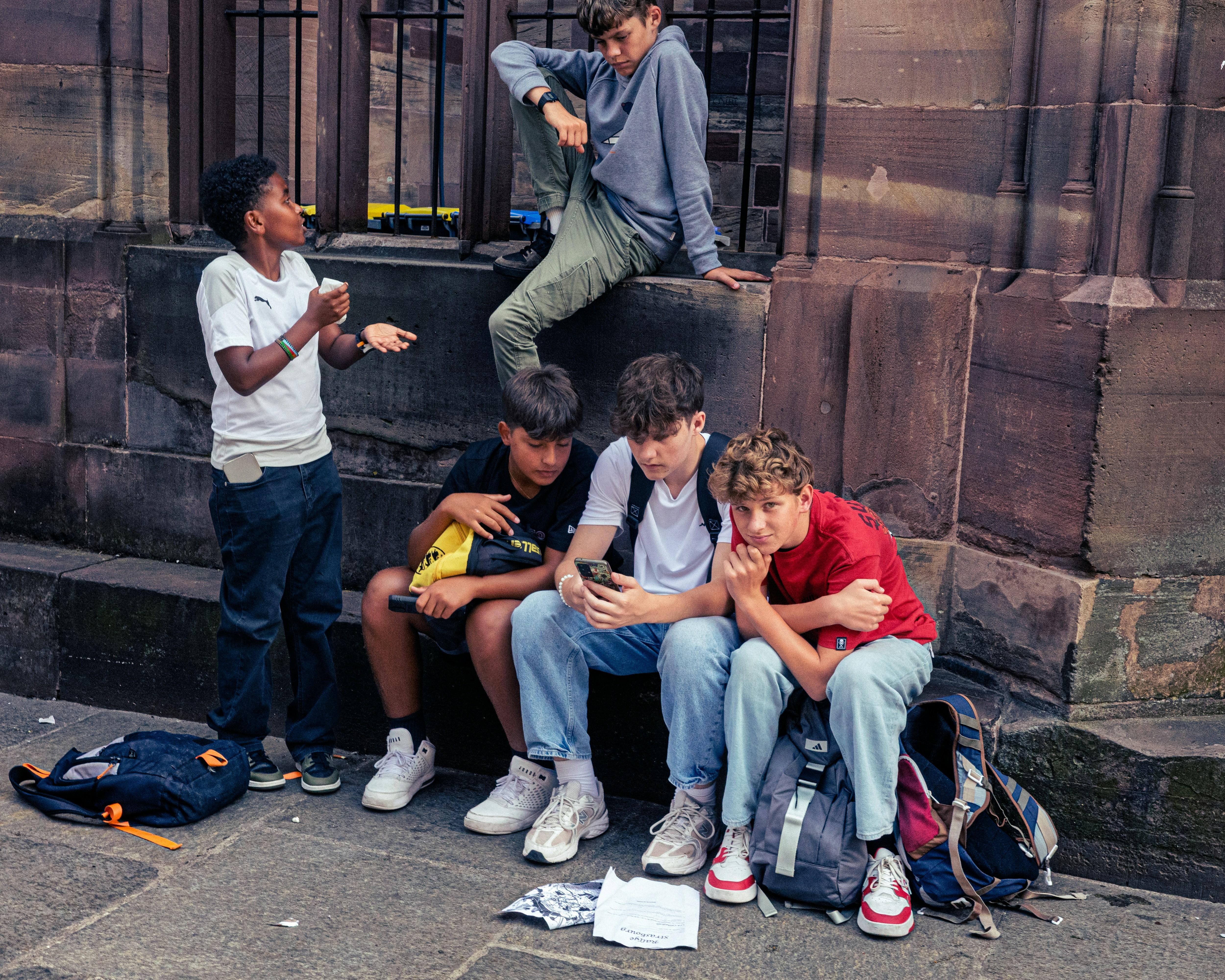 Five teenage boys. Three are sitting on the kerb, one is standing up and the other is sitting on a ledge. They are using their phones and chatting.