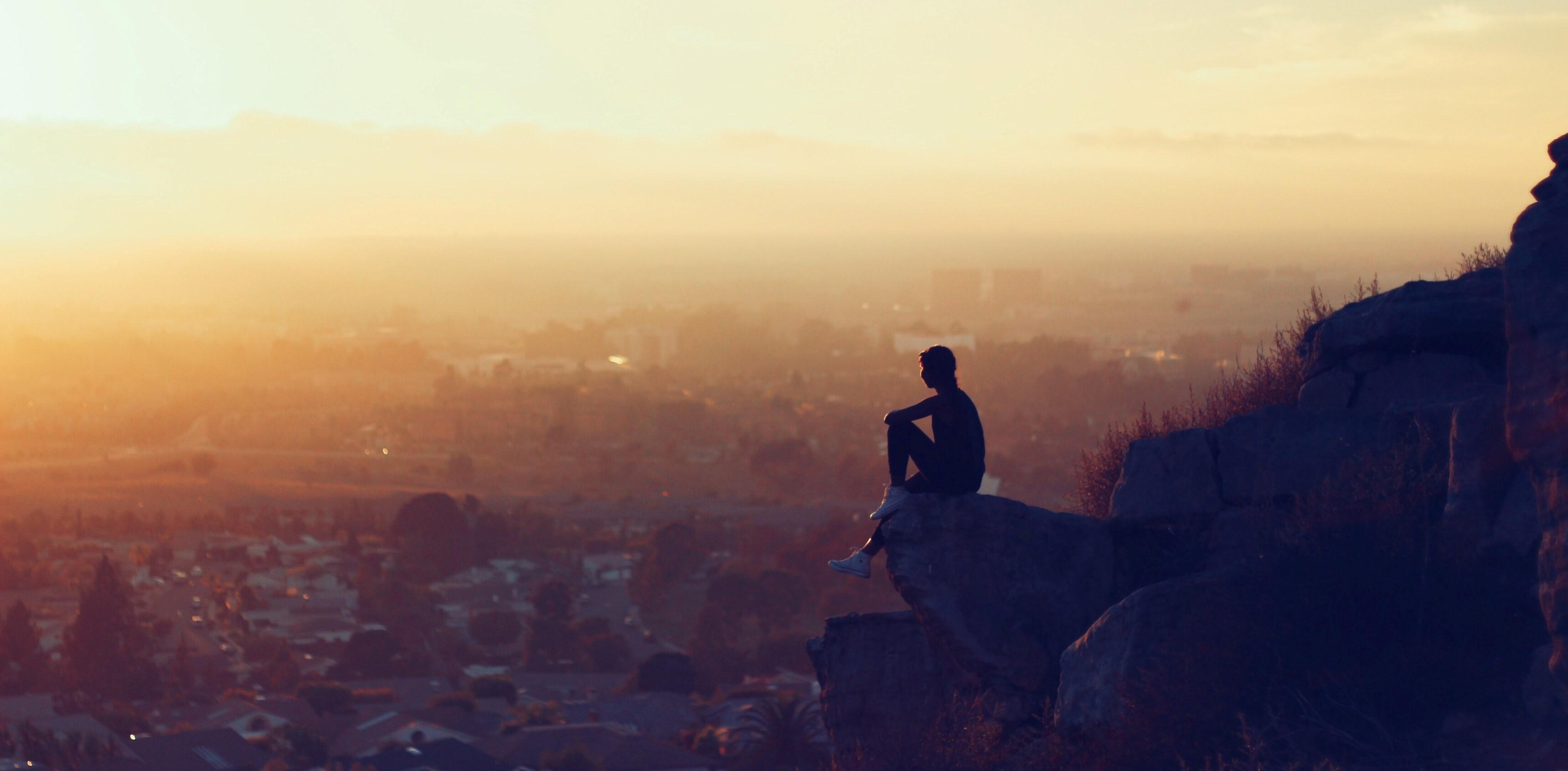 a man sitting on the edge of a cliff looking out on the sunset