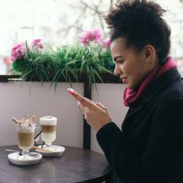 two young women sitting at a cafe having coffee, the one on the left is taking a picture of the coffees and the one on the right is looking at her phone