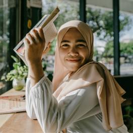 young woman sitting in a cafe holding a book and smiling at the camera