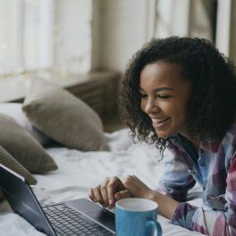 young girl lying on her bed typing on her laptop and smiling