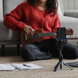 a young woman holding a ukulele sitting on the floor filming herself with a phone in a tripod