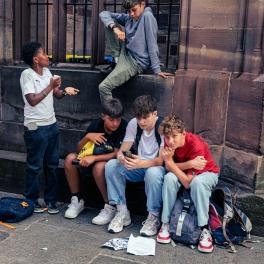 Five teenage boys. Three are sitting on the kerb, one is standing up and the other is sitting on a ledge. They are using their phones and chatting.
