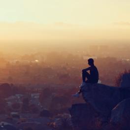a man sitting on the edge of a cliff looking out on the sunset