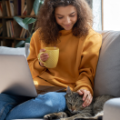 Young woman sitting on the couch with her laptop and a mug, petting a cat