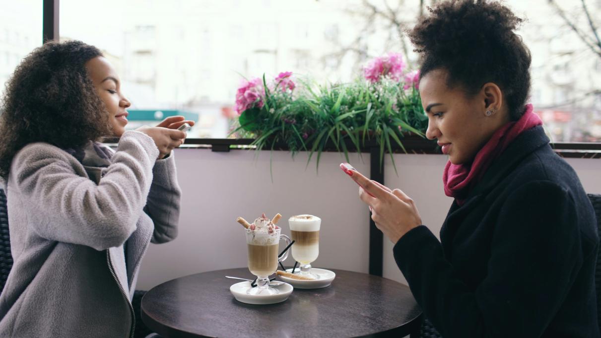 two young women sitting at a cafe having coffee, the one on the left is taking a picture of the coffees and the one on the right is looking at her phone