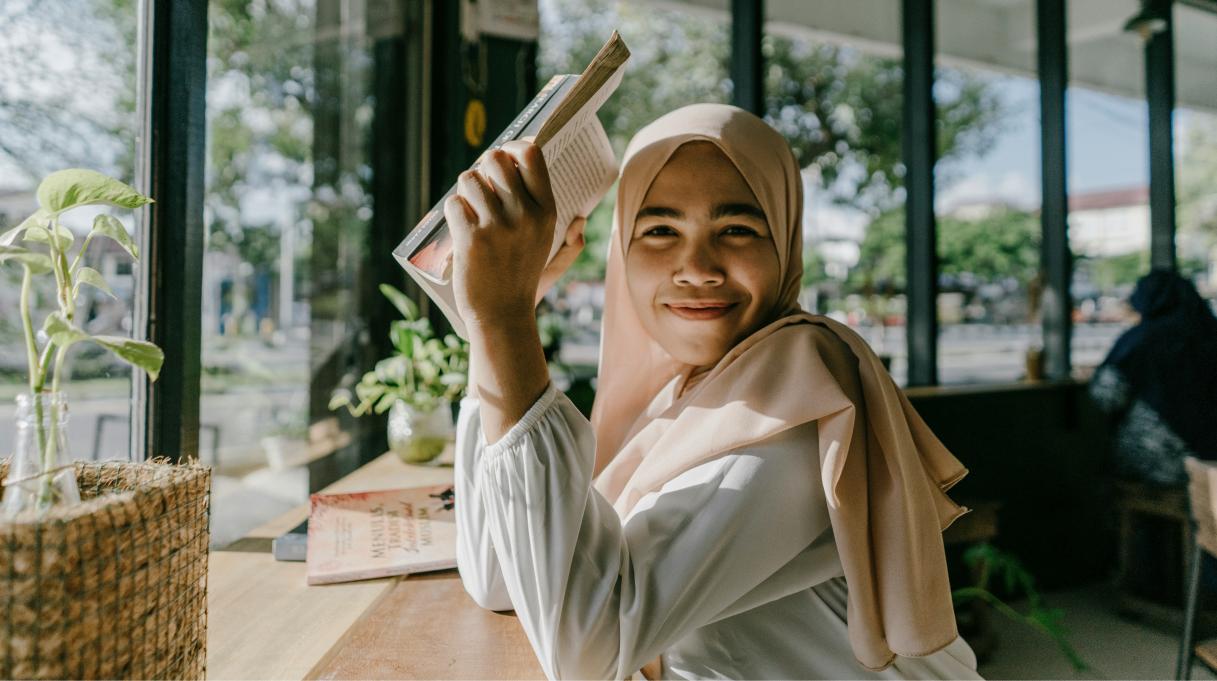 young woman sitting in a cafe holding a book and smiling at the camera