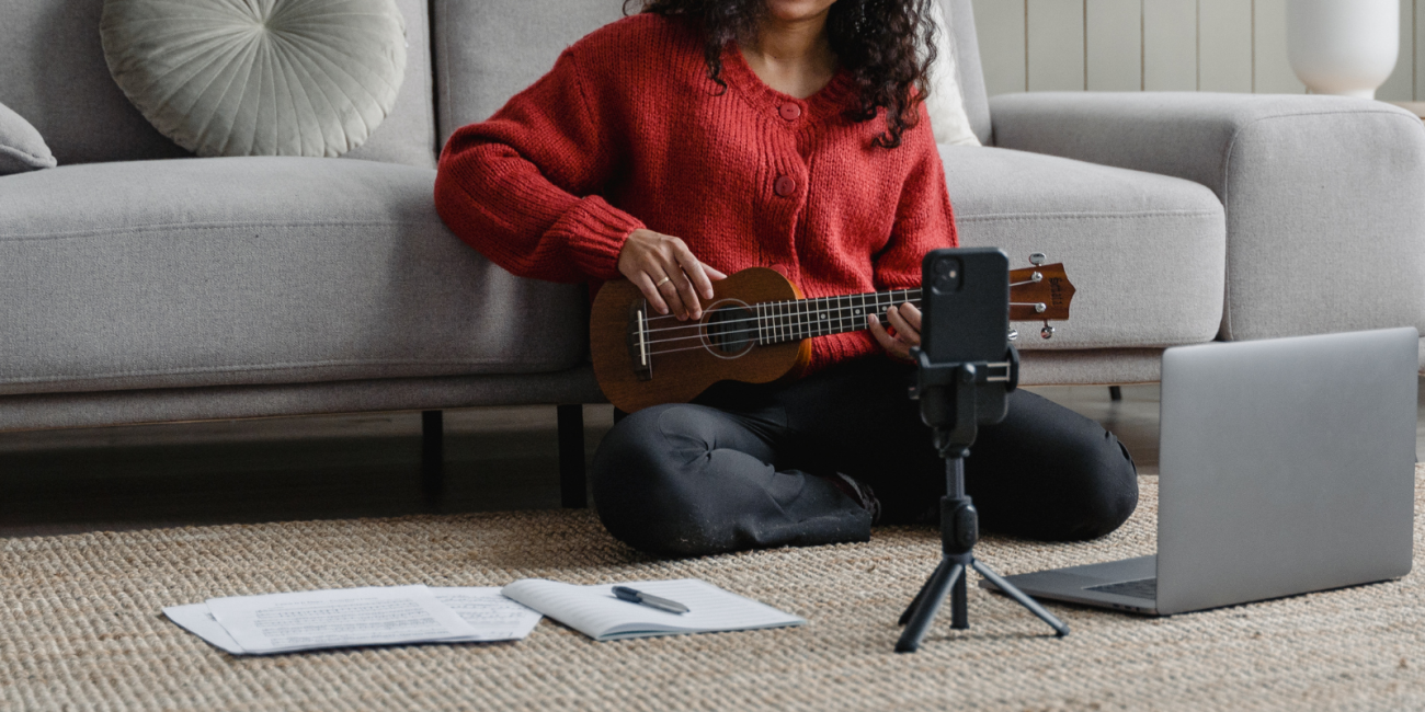 a young woman holding a ukulele sitting on the floor filming herself with a phone in a tripod