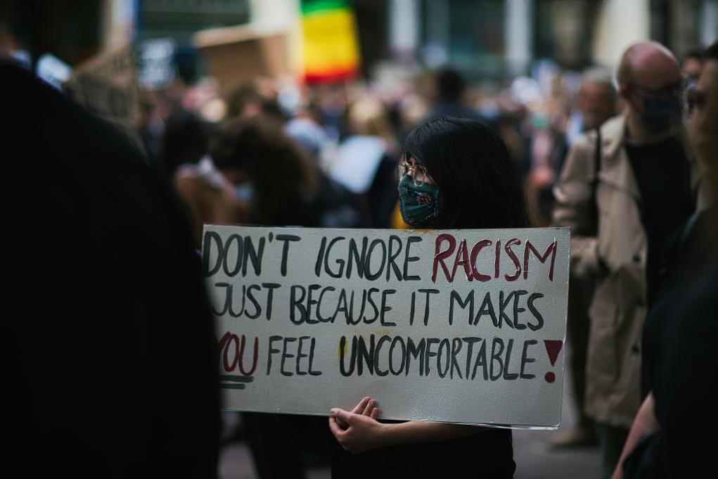 A woman in a mask holding a sign that reads 'Don't ignore racism just because it makes you feel uncomfortable'