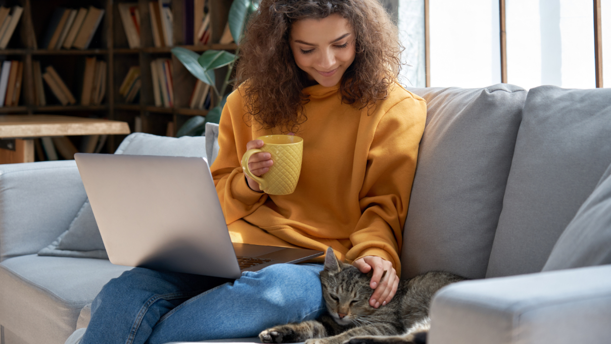 Young woman sitting on the couch with her laptop and a mug, petting a cat